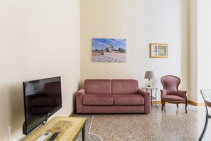 A living room with a flat-screen TV, a pink sofa, a small table with a lamp, and a framed picture on the wall. - Elegant apartment of 66 sqm can accommodate up to 4 people, located on the third floor of a period building with a lift (4 steps to access). The property is located in the historic center of Livorno, just 3 minutes walk from the Duomo and a short walk fr (Livorno)