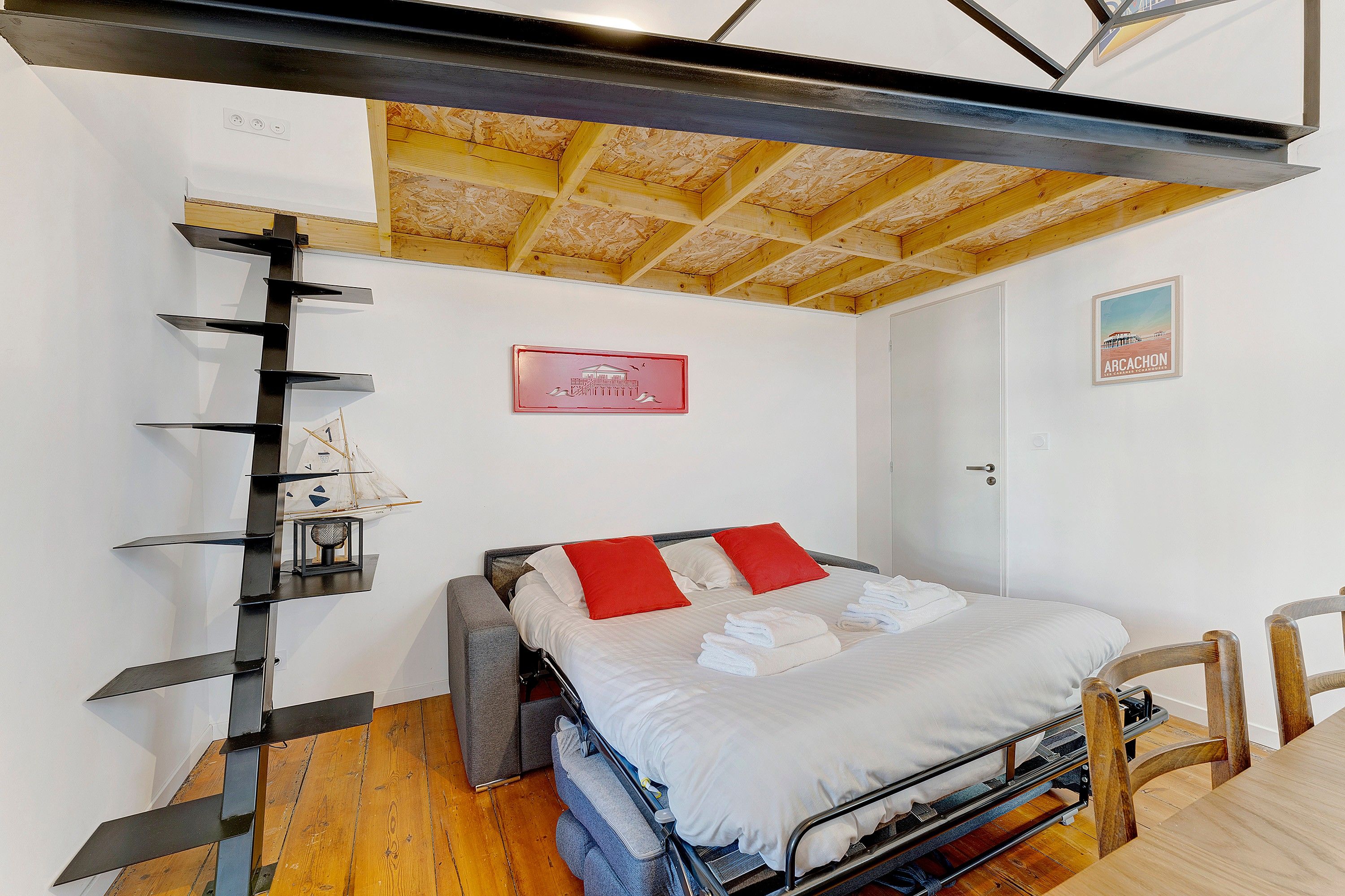 A bedroom with a bed, a wooden floor, a ceiling with exposed beams, a door, and a poster on the wall.