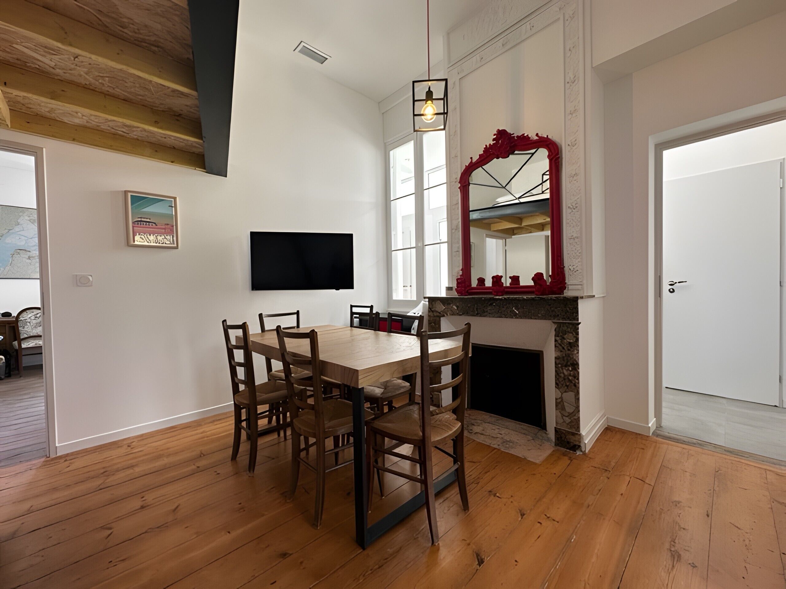A dining area with a wooden table and chairs, a flat-screen TV, a fireplace with a red mirror above it, and a door to the right.
