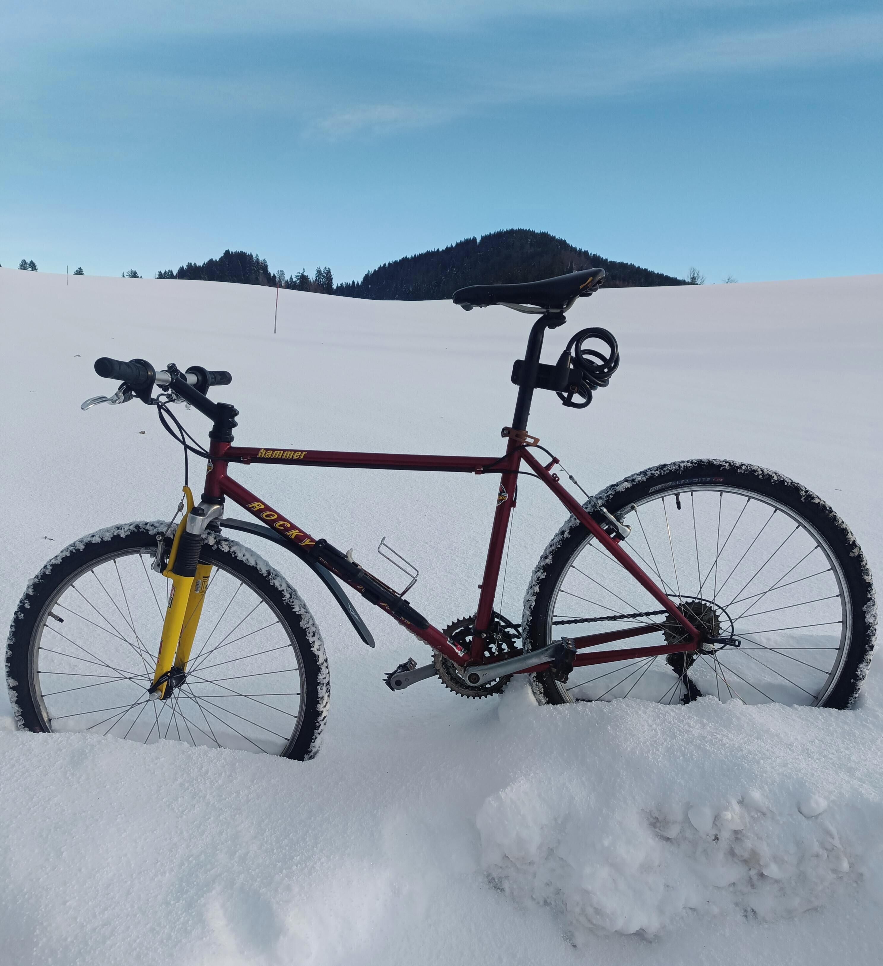 Ein Mountainbike mit rotem Rahmen und gelber Gabel steht in einem verschneiten Feld unter blauem Himmel, im Hintergrund ist eine sanfte Hügelkulisse zu sehen.