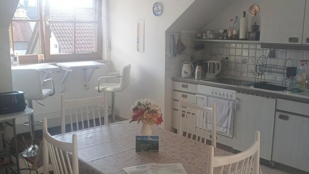 Une cuisine avec des meubles blancs, une table à manger avec des chaises, une fenêtre avec des stores et un ventilateur de plafond.
