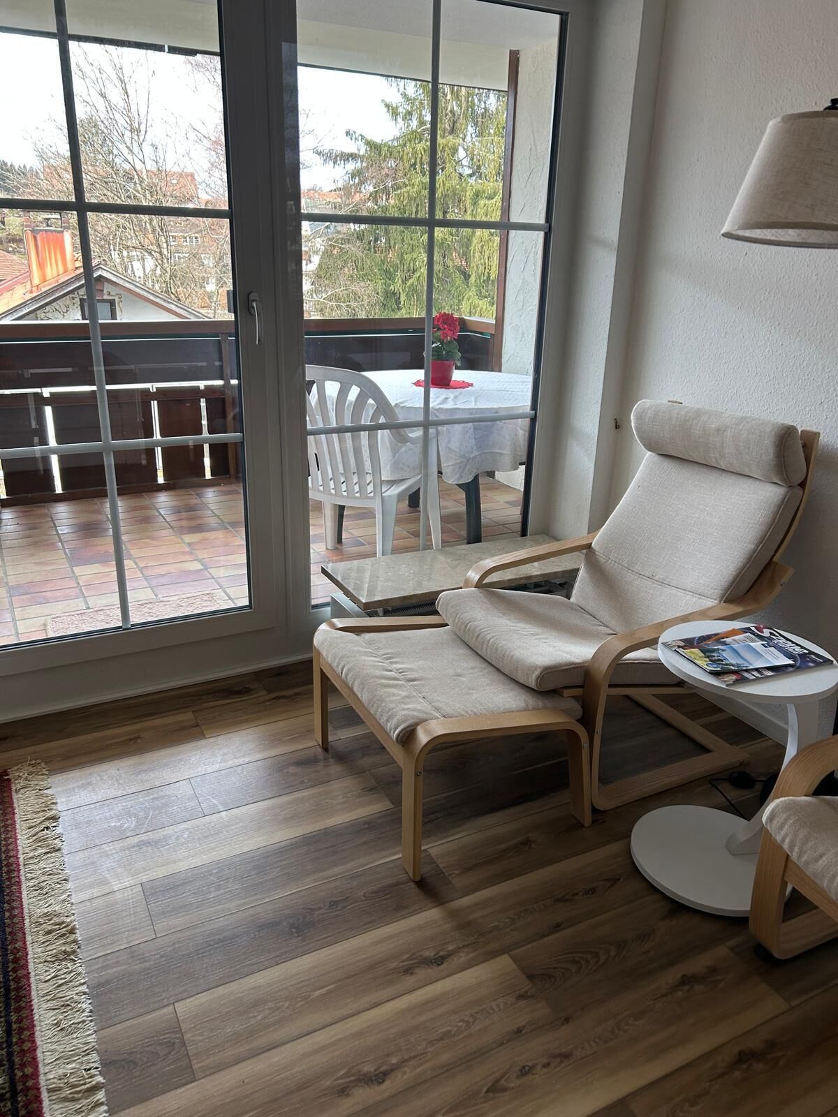 Ein Zimmer mit einer Schiebetür zu einer Terrasse, ein weißer Stuhl, ein kleiner runder Tisch mit einer Zeitschrift und ein Holzboden.