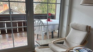 Une pièce dotée d’une porte-fenêtre coulissante donnant sur un patio, d’une chaise blanche, d’une petite table ronde avec un magazine, et d’un plancher en bois.