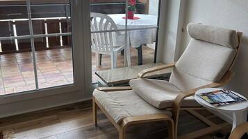 Ein Zimmer mit einer Schiebetür zu einer Terrasse, ein weißer Stuhl, ein kleiner runder Tisch mit einer Zeitschrift und ein Holzboden.