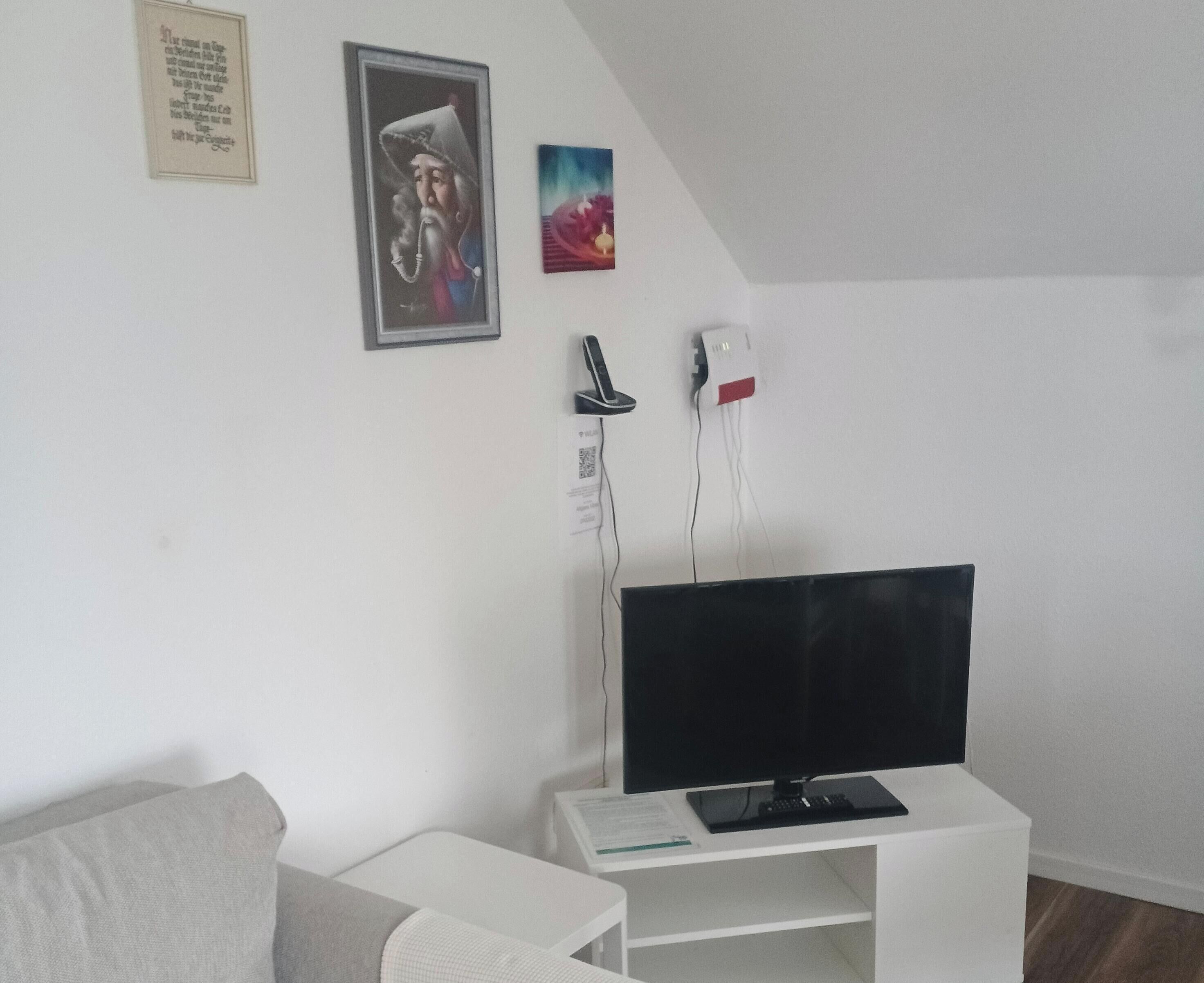 A room with a white sofa, a TV on a stand, a framed picture, and a wall-mounted phone.