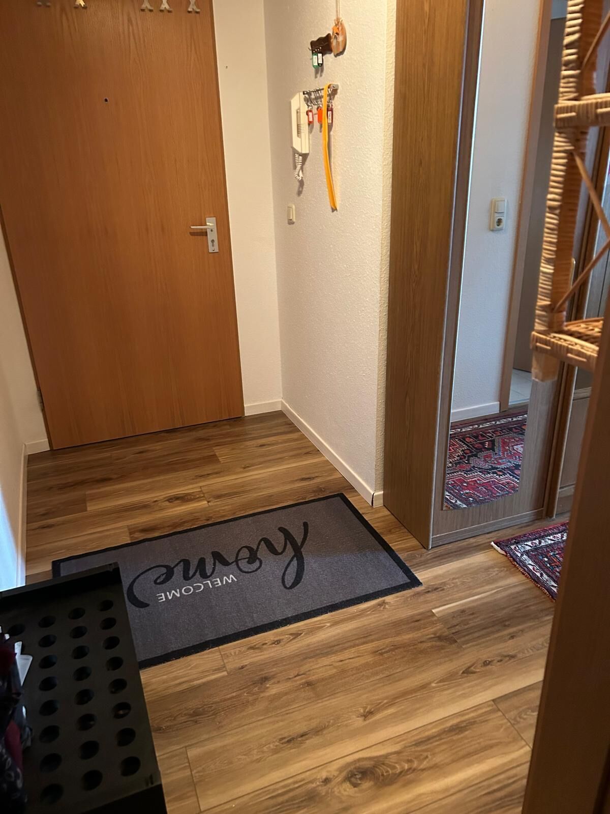 Eine Holztür, ein an der Wand befestigtes Stromdosenfeld, eine Fußmatte mit dem Text 'Welcome' und ein gemustertes Teppich.