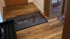 Une porte en bois, un tableau électrique fixé au mur, un paillasson avec l’inscription « Bienvenue » et un tapis à motifs.