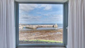 Une pièce dotée d’une grande fenêtre donnant sur la côte, avec une digue en béton et un phare visibles.
