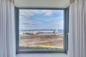 A room with a large window overlooking a coastal area, featuring a concrete breakwater and a lighthouse. - Seal`s Bay Luxury Holiday Home for 4 in Seahouses (Northumberland)