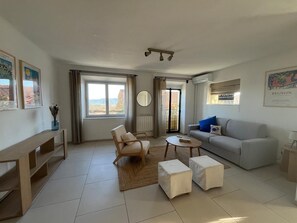 A living room with a sofa, armchair, coffee table, and ottomans. There is a window with curtains, a radiator, and a wall-mounted air conditioning unit. - 1900 traditional house in Grimaud (Grimaud)