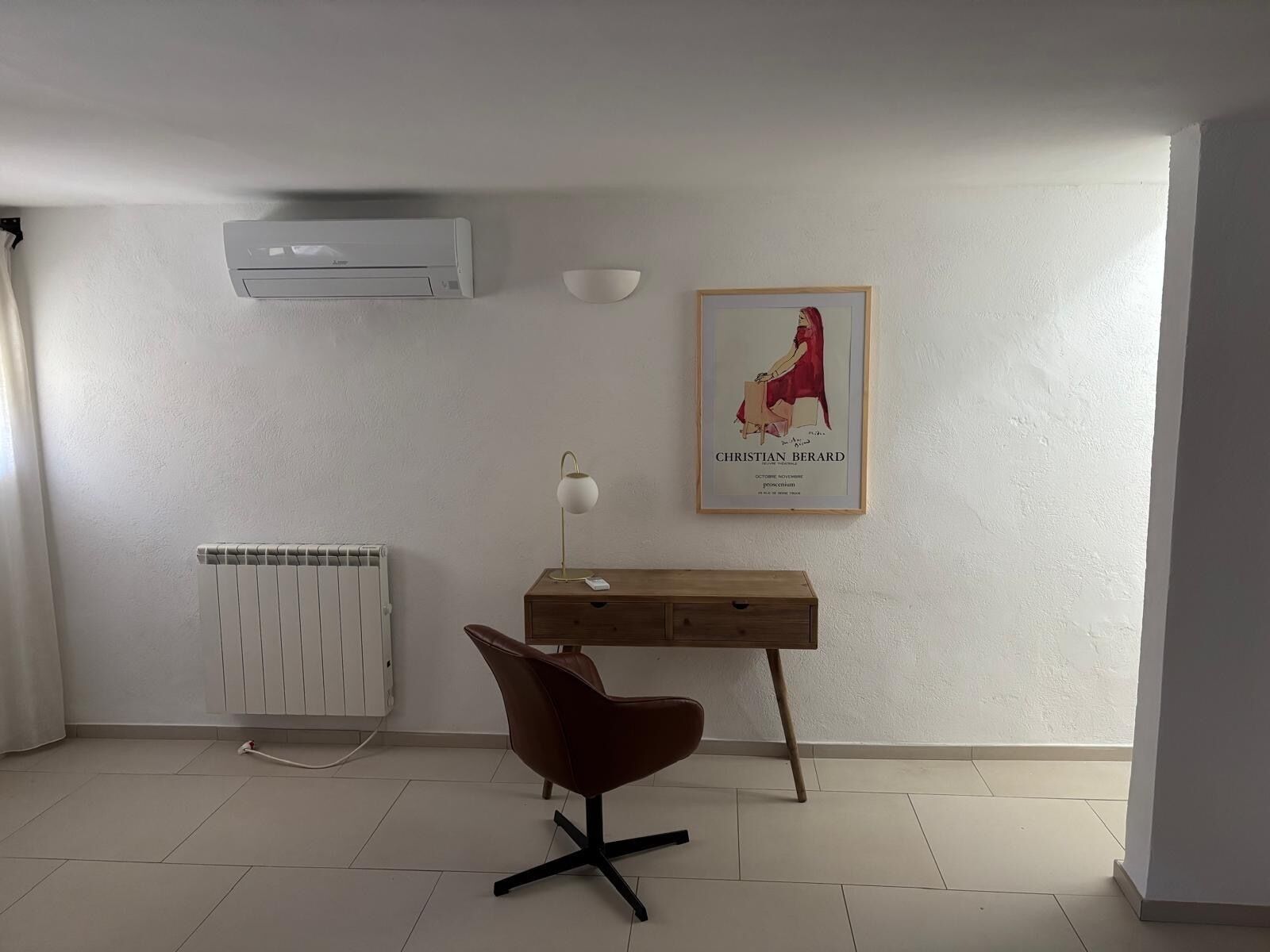Ein Zimmer mit Heizkörper, Klimaanlage, Schreibtisch, Stuhl, Lampe und einem gerahmten Bild an der Wand.