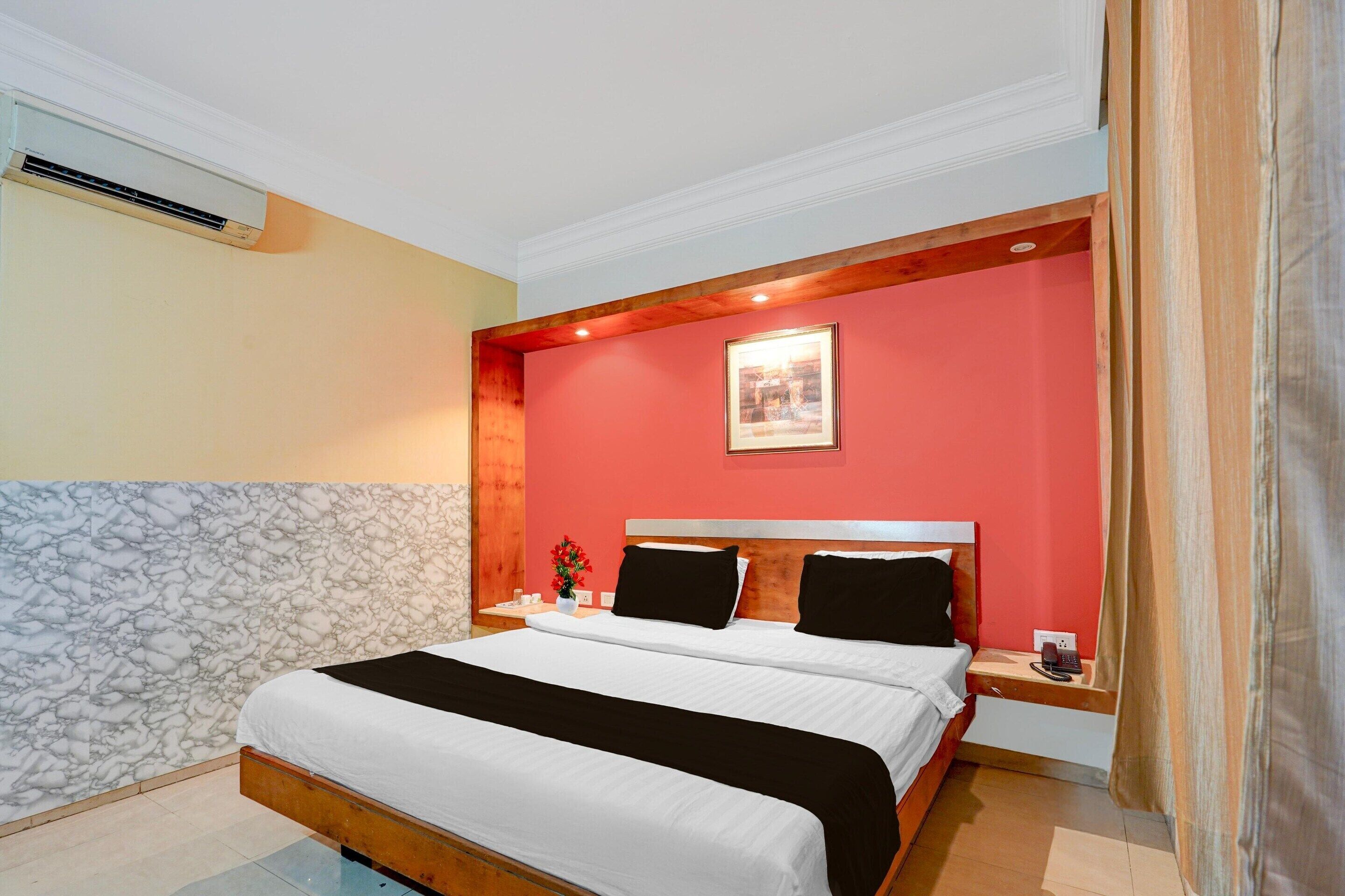A hotel room with a bed, a nightstand, an air conditioning unit, a framed picture on the wall, and a window with curtains.