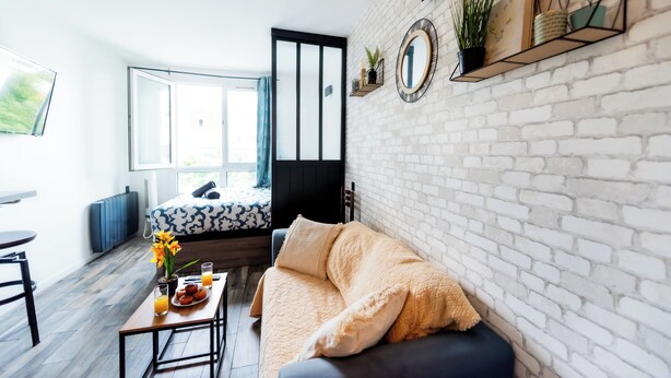 En moderne stue med sofa, sofabord og blomsterarrangement, samt seng med blå og hvidt mønster på dyne.