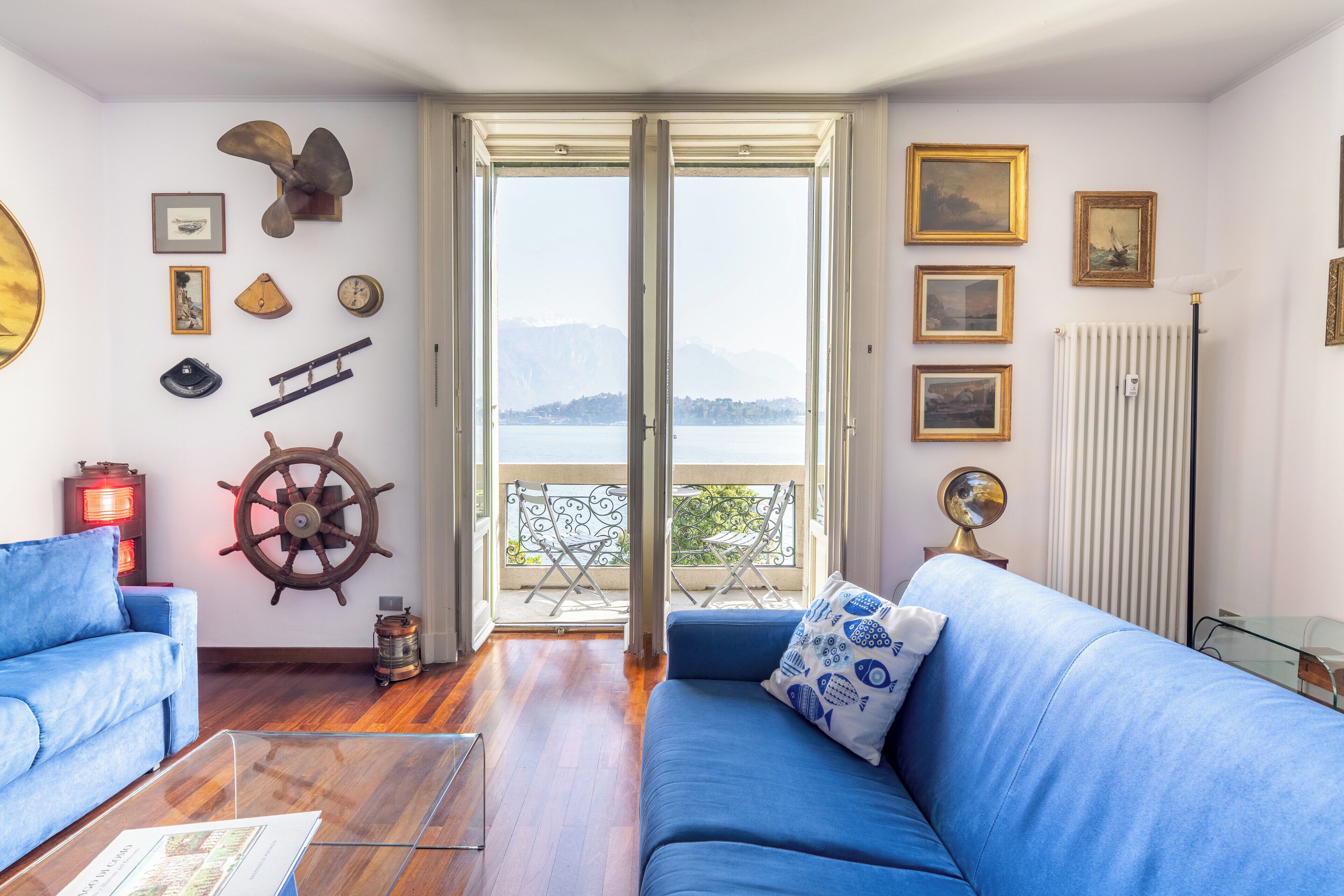 Une pièce avec un canapé bleu, un plancher en bois, un élément décoratif en forme de roue de navire et une vue sur la mer par une grande fenêtre.