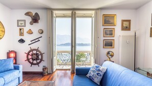 A room with a blue sofa, a wooden floor, a ship's wheel decoration, and a view of the sea through a large window. - Miraluna – Lake Como View Balcony (Griante)