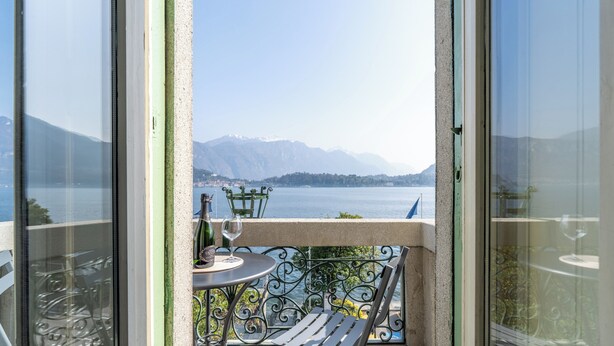 A balcony with a table, chair, and wine glass, overlooking a lake and mountains. - Miraluna – Lake Como View Balcony (Griante)