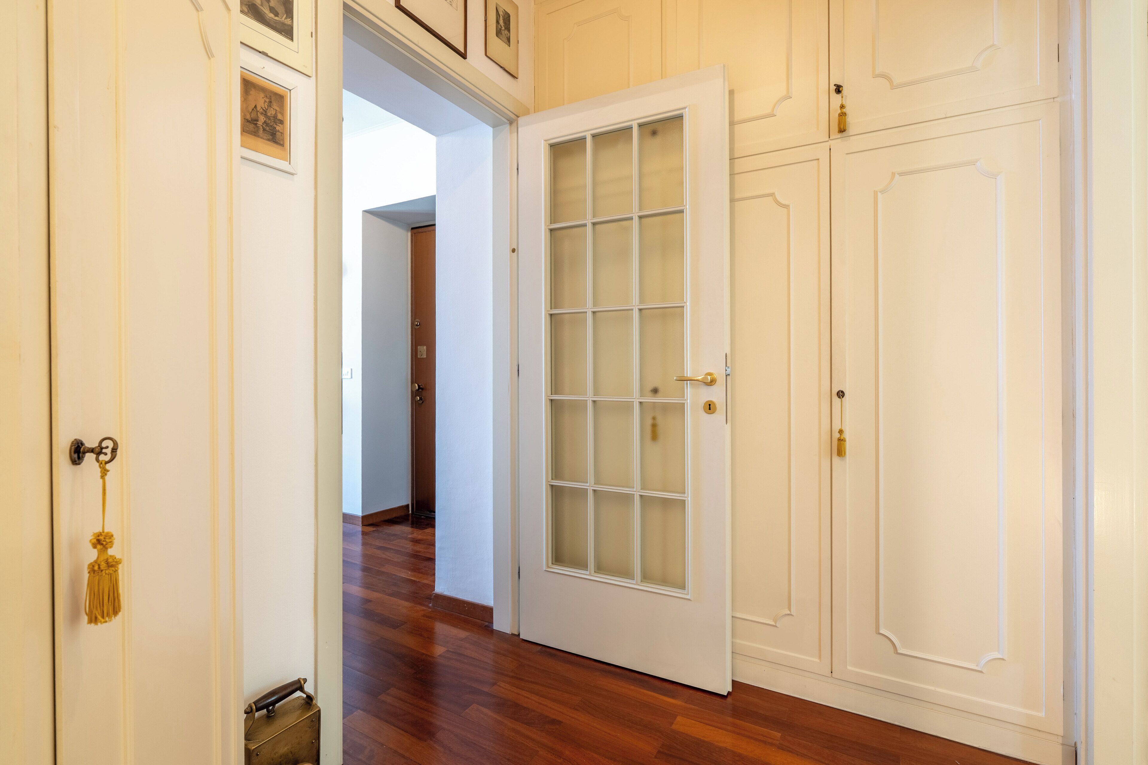 Un couloir avec une porte blanche dotée de panneaux de verre, un plancher en bois et un mur blanc orné d’un cadre.