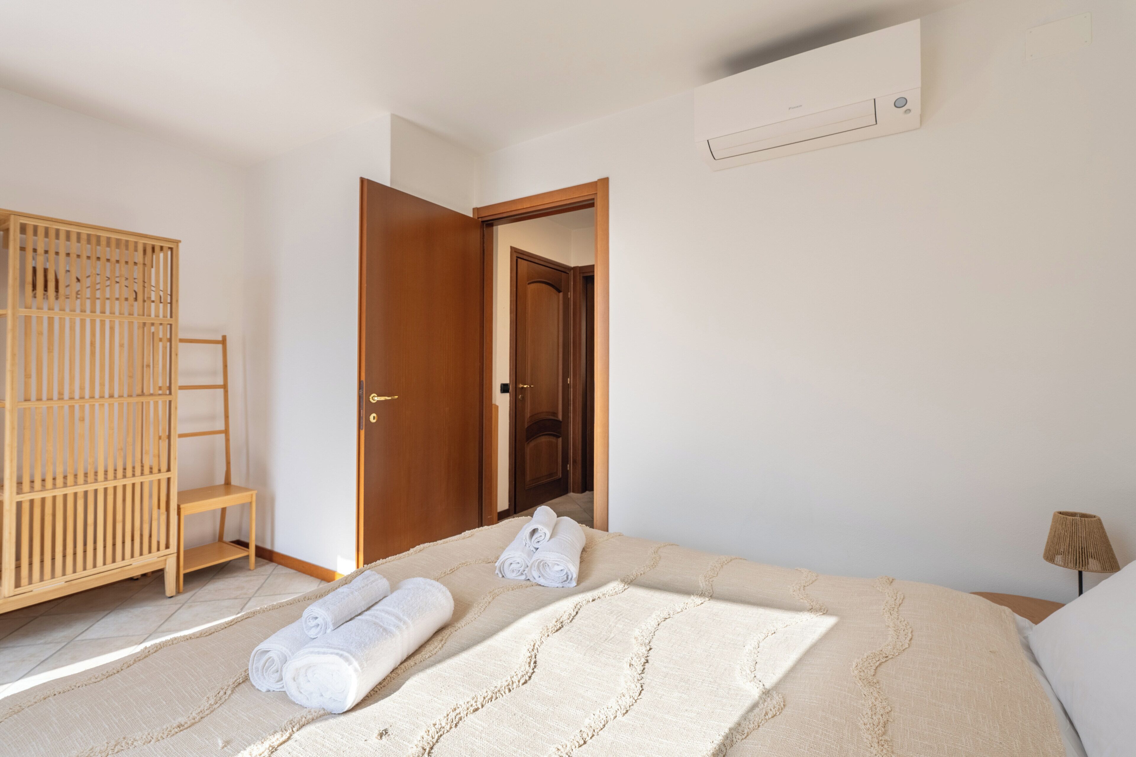 A bedroom with a bed, rolled towels, a wooden wardrobe, a door, and a wall-mounted air conditioner.