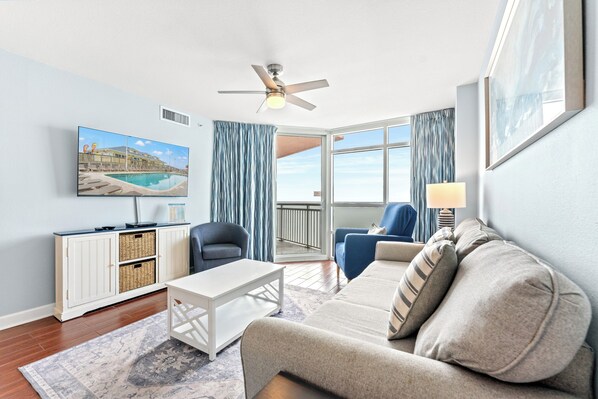 A modern living room with a sofa, armchairs, a coffee table, a TV mounted on the wall, and a ceiling fan. - Prince Resort 1904- Oceanfront 2 Bedroom-Pool (North Myrtle Beach)