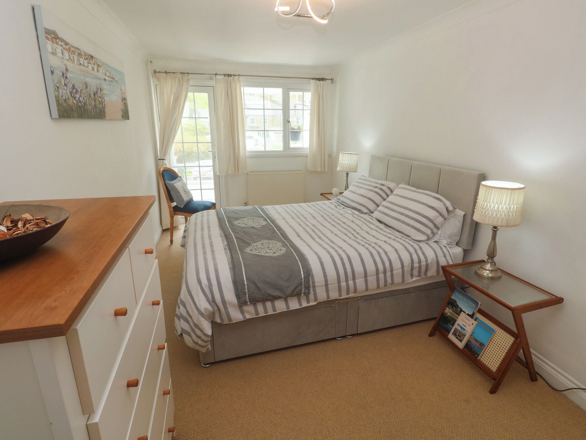 A bedroom with a bed, a chair, a bedside table with a lamp, a window with curtains, and a painting on the wall.