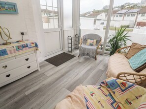 A room with a wooden floor, a white dresser with heart-shaped drawers, a wicker chair, a sofa, and a view of houses through large windows. - 16 Forth An Nance (Redruth)