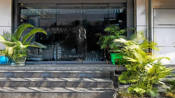 A building entrance with a glass door, steps leading up to it, and potted plants on either side. - Deluxe Hotel Okasu Railway Station (Raipur)