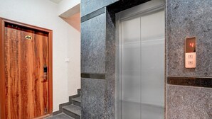 A wooden door with a number plate, a marble wall, and an elevator with a digital display. - Classic With Balconyroyal Avenue (Bengaluru)