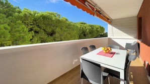A balcony with a table and chairs, a red and white awning, and a view of green trees. - Modern Seaside Flat With Spacious Balcony (Lignano Sabbiadoro)