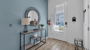 Un pasillo con pared azul, un espejo redondo, una consola con lámpara, una planta pequeña y un extintor.