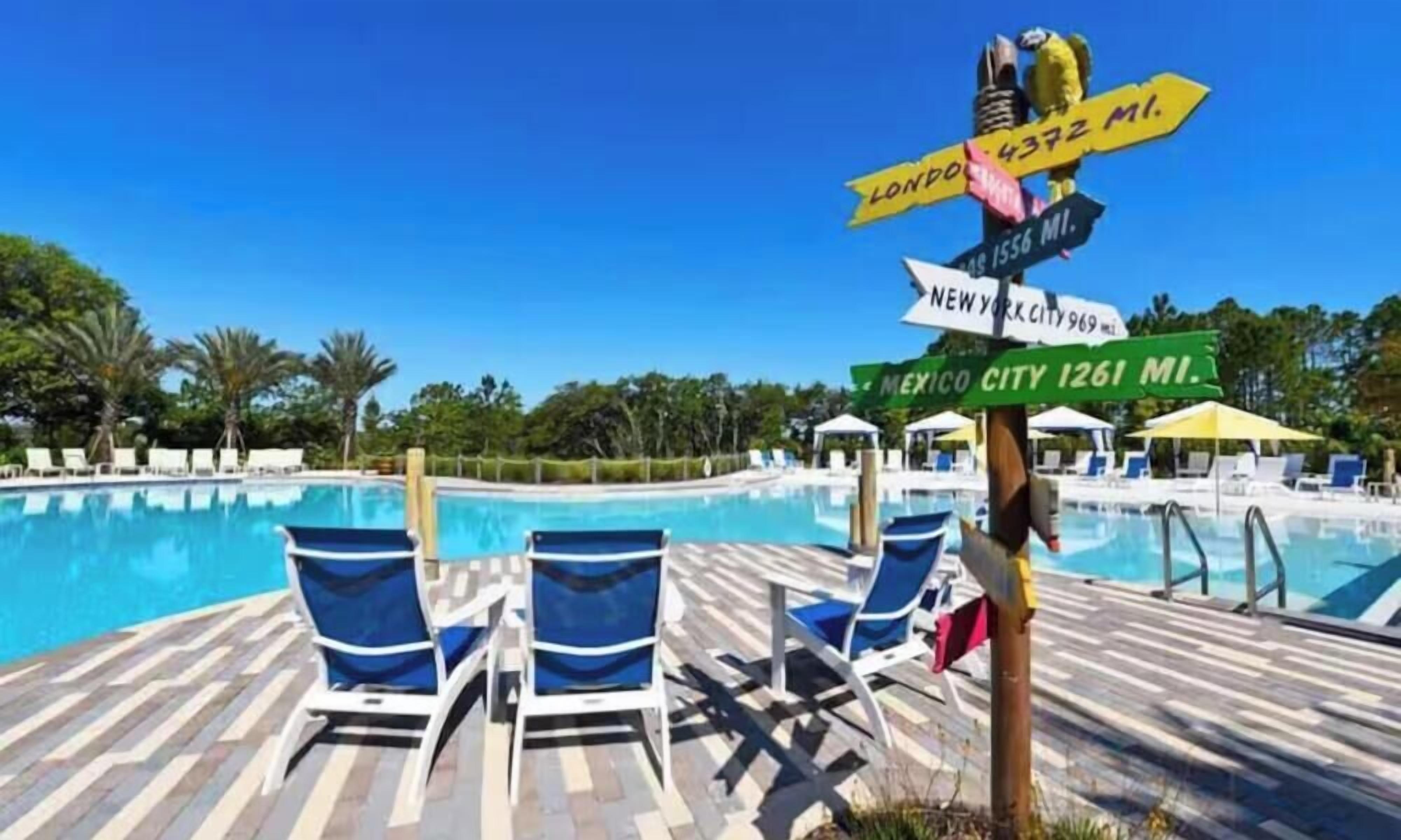 Un espace aménagé au bord de la piscine, avec des chaises longues, un panneau indiquant les distances vers les différentes villes, et un ciel d’un bleu limpide.