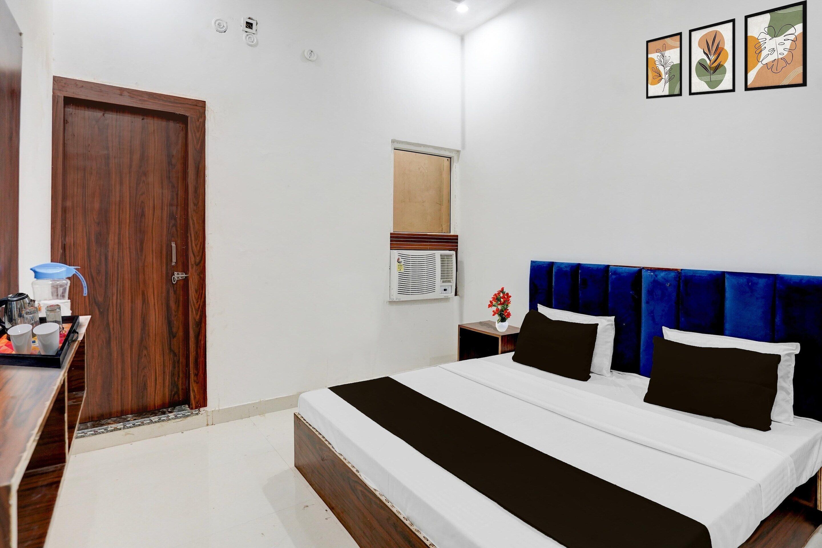 A bedroom with a wooden door, a bed with a blue headboard, a bedside table with a lamp, and a wall-mounted air conditioner.