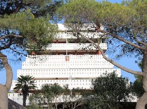 Um edifício de vários andares com fachada branca, varandas e rodeado por árvores.