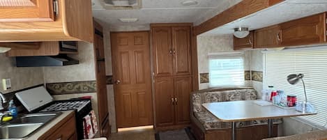 Cozinha compacta de motorhome com armários de madeira, fogão, pia e área de refeições com mesa e banco.