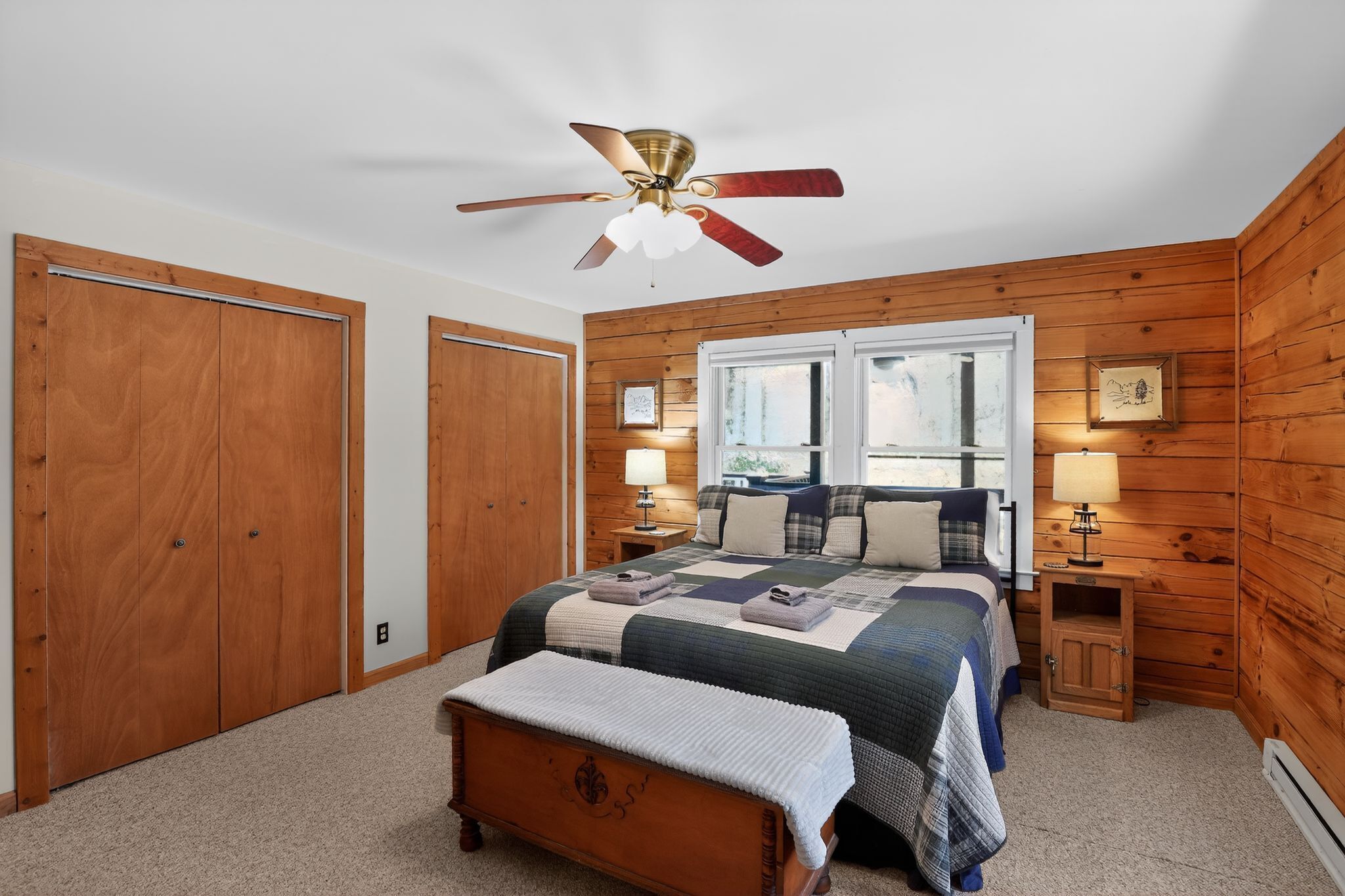 A bedroom with a bed, wooden walls, a ceiling fan, two bedside lamps, a window, and a wooden bench.