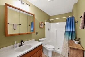 A bathroom with a wooden vanity, a white sink, a toilet, a shower with a blue curtain, and a mirror with lights. - Blue Ridge Retreat w/ Private Trail (Pisgah Forest)