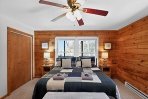 A bedroom with a wooden paneled wall, a bed with a quilt, two bedside tables with lamps, and a ceiling fan. - Blue Ridge Retreat w/ Private Trail (Pisgah Forest)