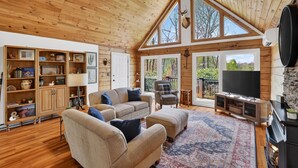 A spacious living room with wooden walls and ceiling, a large window, a fireplace, and a television. - Blue Ridge Retreat w/ Private Trail (Pisgah Forest)