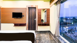 A hotel room with a bed, a TV mounted on the wall, a wooden door, a mirror, and a window with a city view.