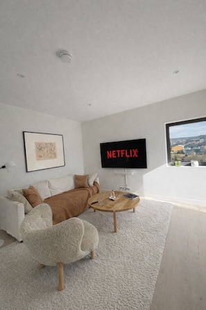 Een moderne woonkamer met een witte bank, een ronde salontafel, een groot televisiescherm waarop het Netflix-logo staat en een uitzicht op een woonwijk door een raam.