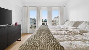 Un dormitorio con cama, televisor, cómoda y vistas a edificios a través de amplias ventanas.