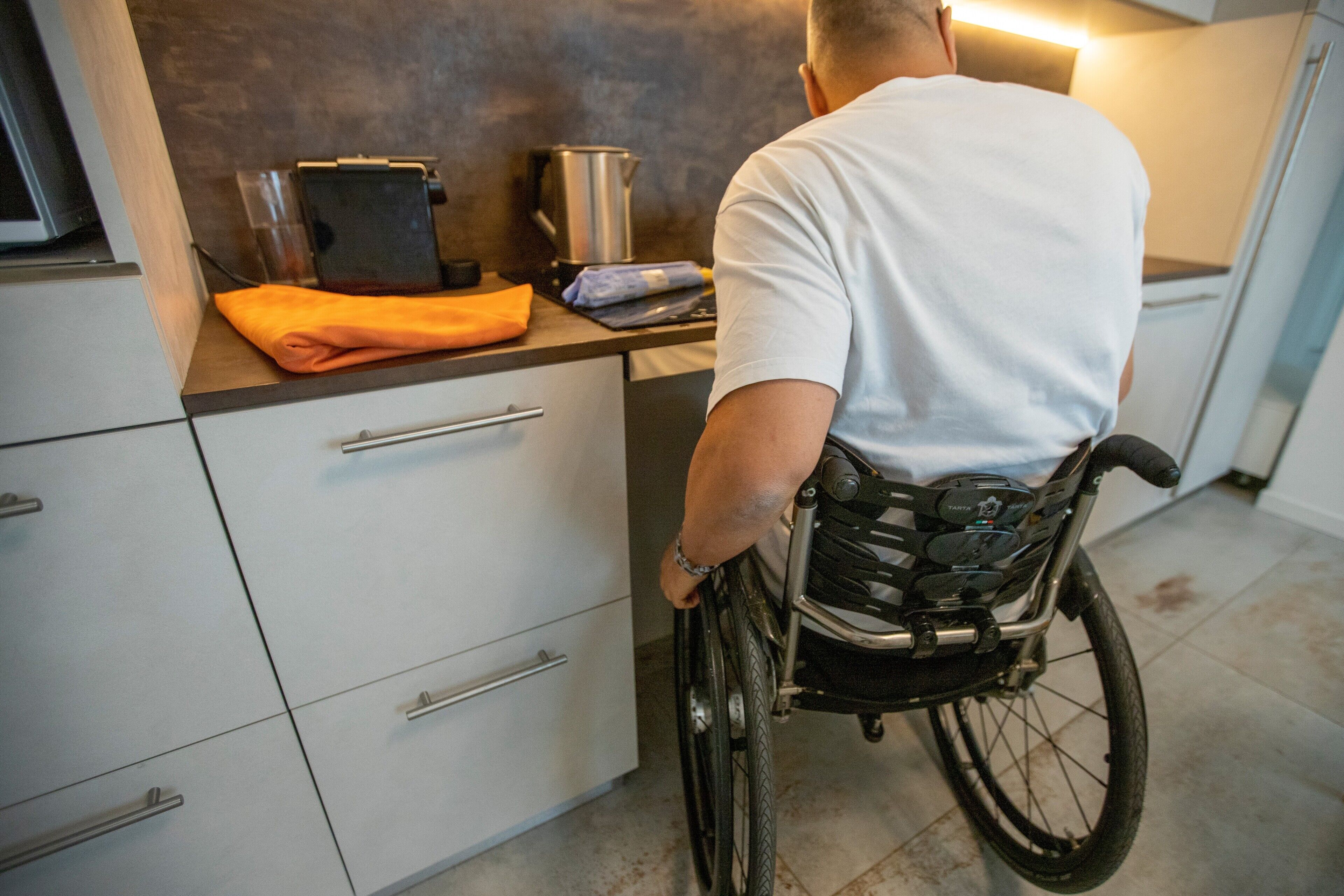 A person in a wheelchair in a kitchen with cabinets, a countertop, and a kettle.