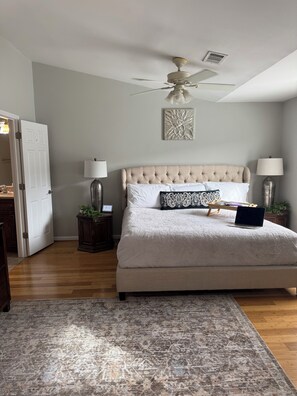 Un dormitorio con cama, ventilador de techo, mesita de noche con lámpara, alfombra y puerta que da a otra habitación.