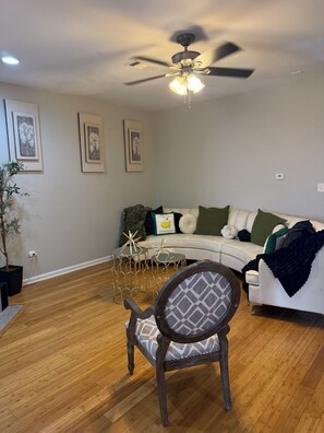 Una sala de estar con ventilador de techo, un sofá blanco con cojines verdes y blancos, un sillón estampado y una mesita con objetos decorativos.
