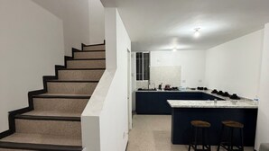 Una cuina moderna amb una illa central i tamborets, una escala amb baranes negres, i un sofà gris.