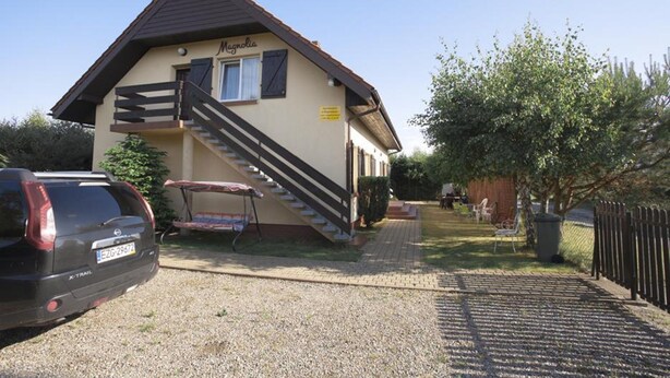 Ein zweistöckiges Gebäude mit einer Treppe, einem geparkten Auto davor und einem von einer Hecke umgebenen Bereich mit Bäumen.