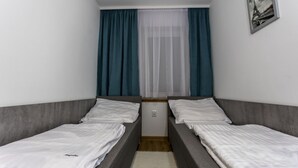 Ein kleiner, schmaler Raum mit zwei Einzelbetten, einem Fenster mit durchsichtigen Vorhängen und einem gerahmten Bild an der Wand.