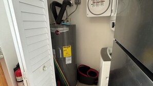 Eine kleine Küche mit Kühlschrank, Durchlauferhitzer, Besen und Feuerlöscher.