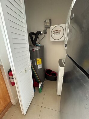 A compact kitchen with a refrigerator, a water heater, a broom, and a fire extinguisher. - Delightful 1-bedroom apartment with AC in vibrant Wellington (Wellington)
