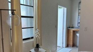 Ein Schlafzimmer mit einem Bett, einem kleinen weißen Hocker, einer Topf-Orchidee, einer Stehlampe und Blick auf eine Küchenzeile.