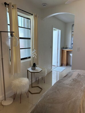 A bedroom with a bed, a small white stool, a potted orchid, a floor lamp, and a view of a kitchen area. - Delightful 1-bedroom apartment with AC in vibrant Wellington (Wellington)
